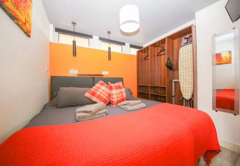 Compact bedroom with red bedding, orange accent wall, wooden wardrobe, and ceiling light