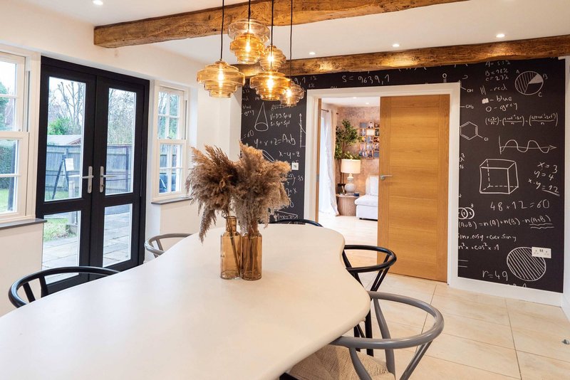 Open-plan kitchen and dining area with white countertop, pendant lights, pampas grass arrangement, and chalkboard wall