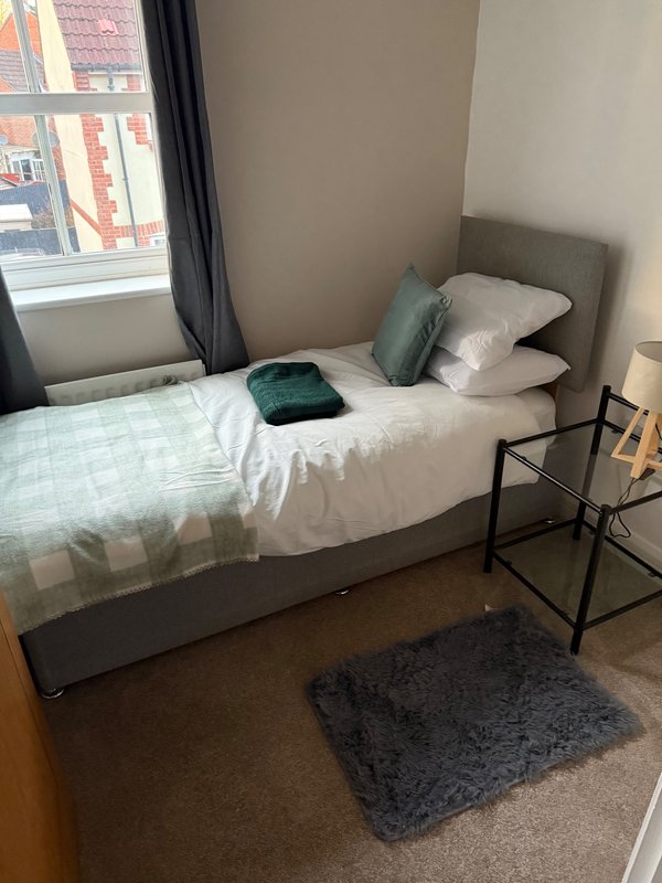 Compact bedroom with single bed, green cushions, window with city view and bedside table