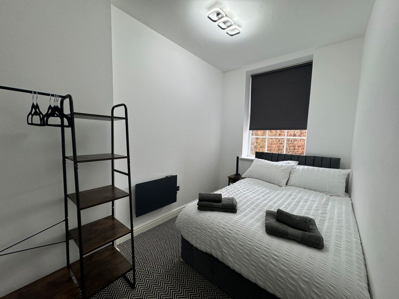 Compact bedroom with double bed, metal shelving unit, dark roller blind and wooden flooring