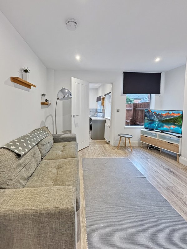 Open-plan living room with beige sofa, TV, wooden floor and access to kitchen and bathroom