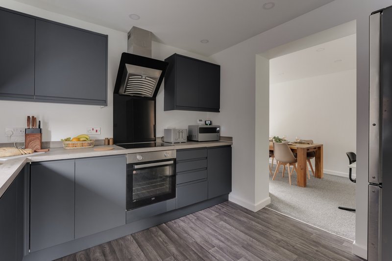 Modern kitchen with dark cabinetry, built-in oven, black kettle, and wooden dining table visible through doorway