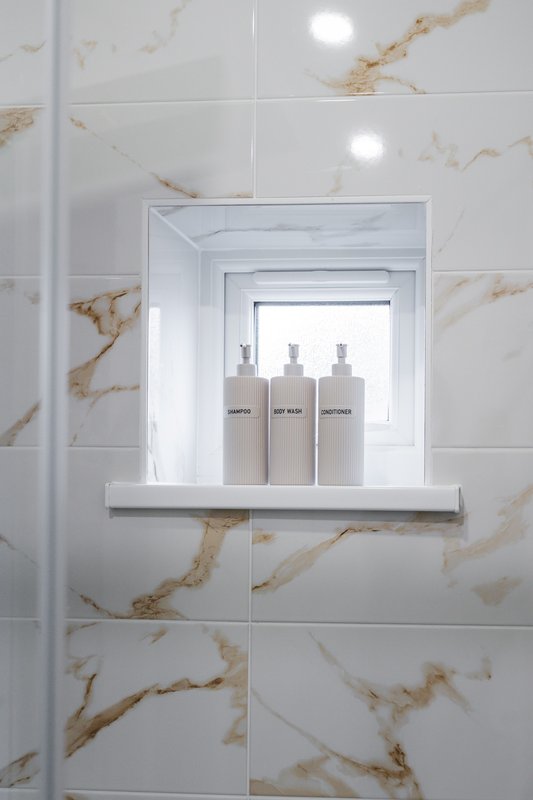 Modern bathroom niche with three white toiletry bottles on white shelf, marble-effect tiling