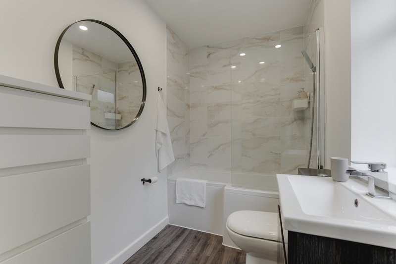 Modern bathroom with marble shower enclosure, round mirror, white vanity and dark flooring
