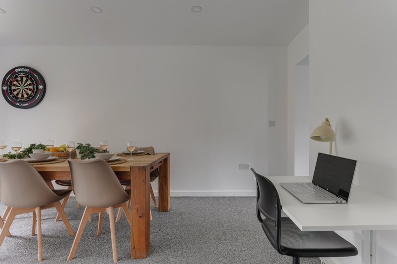 Open-plan workspace with wooden dining table, chairs, desk with laptop, and dartboard on wall