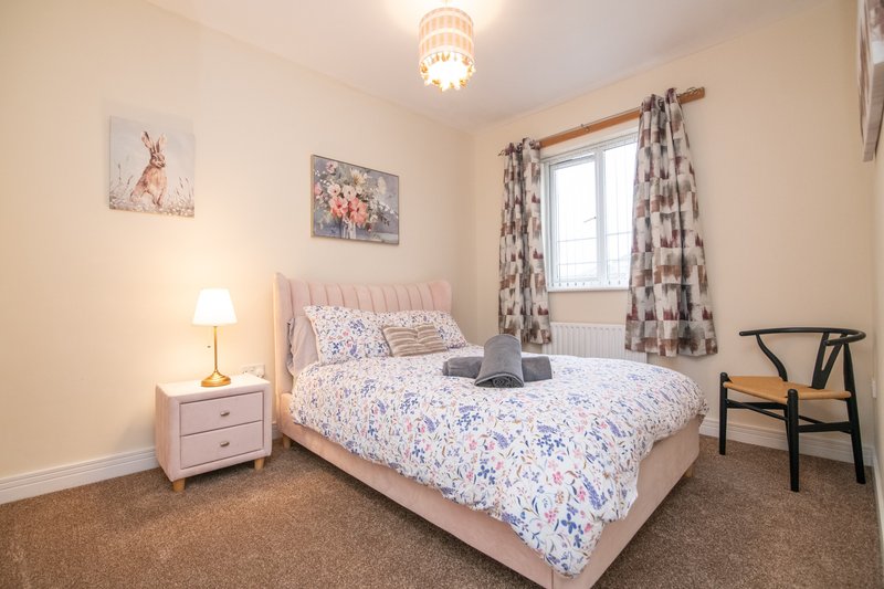 Bright bedroom with pink upholstered bed, bedside table, lamp, window with patterned curtains, and wooden chair