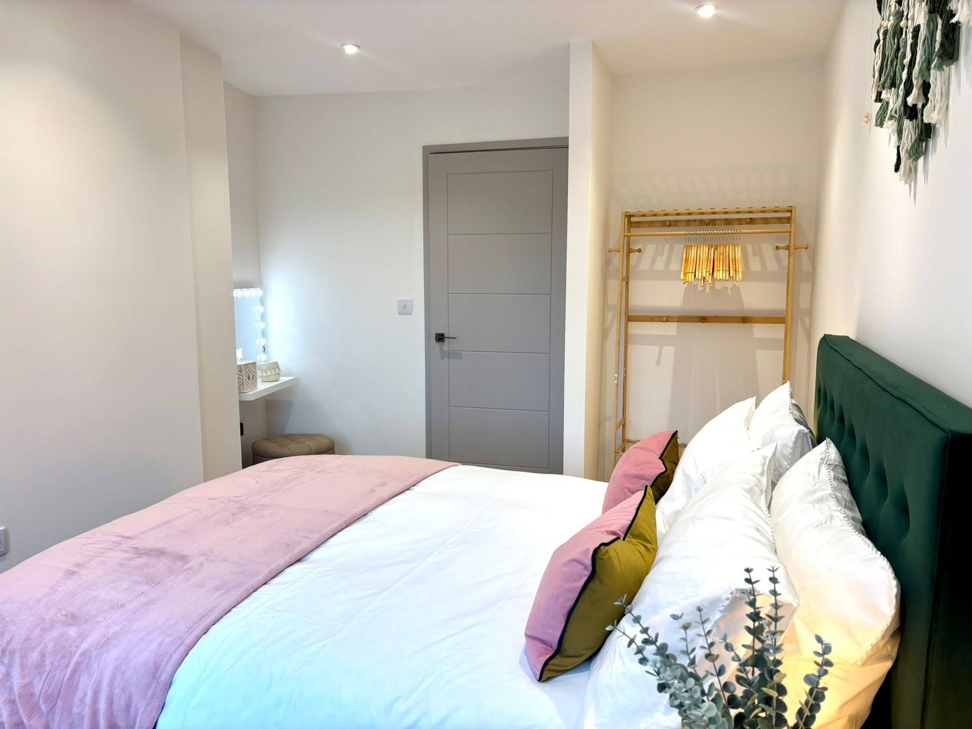 Bedroom with double bed, green headboard, colorful bedding, wooden shelving and grey door