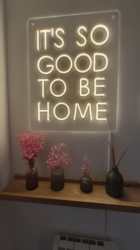 Neon sign reading 'It's So Good to Be Home' above wooden shelf with dried flowers in bottles