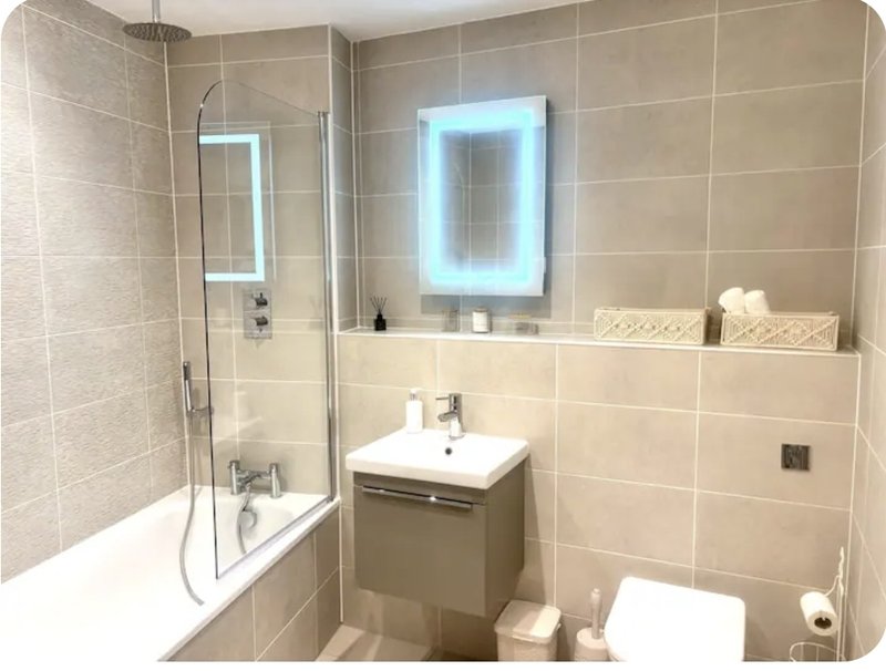 Modern bathroom with beige tiles, shower bath, illuminated mirror cabinet, and vanity sink