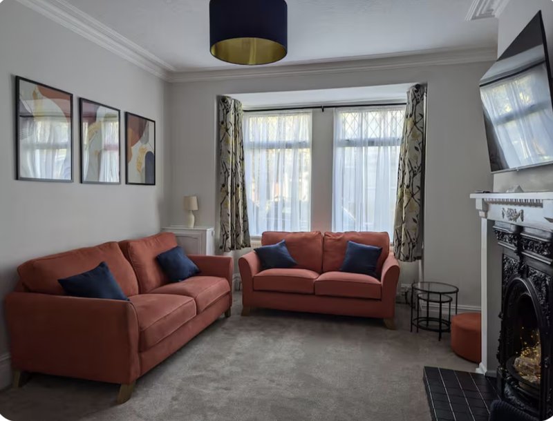Living room with two rust-colored sofas, framed artwork, large windows with curtains, and fireplace