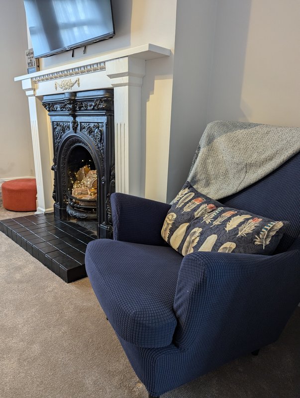 Living room with fireplace, blue armchair, throw blanket, patterned cushion and wall-mounted TV
