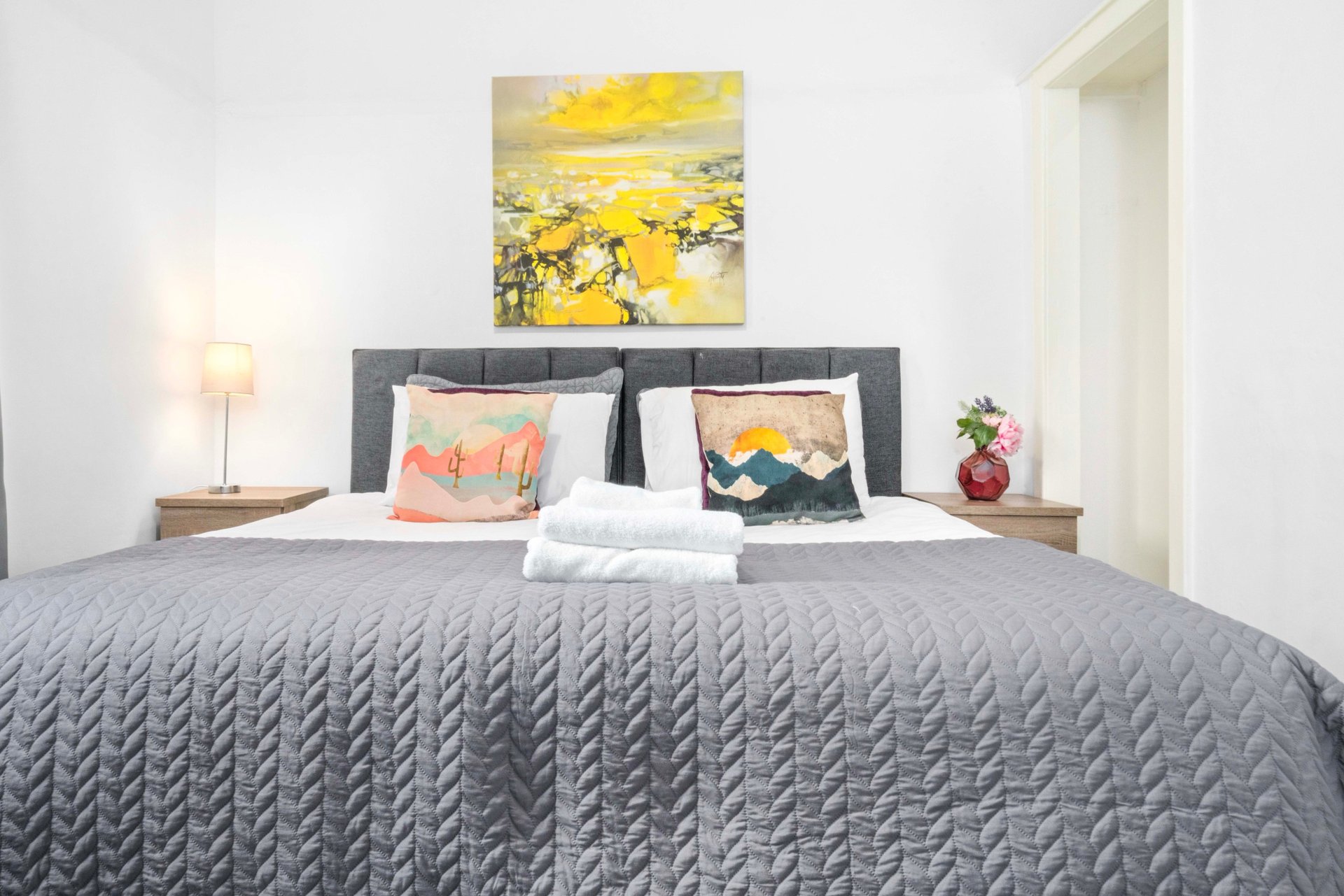 Modern bedroom with gray quilted bed, decorative pillows, framed yellow artwork, and bedside lamp
