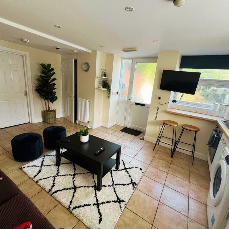 Open-plan living area with black coffee table, blue poufs, patterned rug, TV on wall, and kitchen appliances