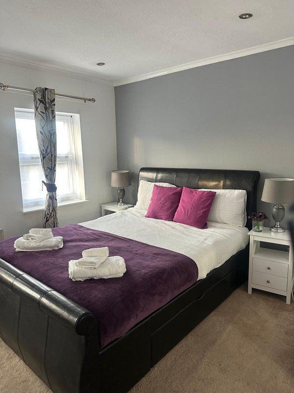 Modern bedroom with double bed, purple throw, pink cushions and white bedside tables