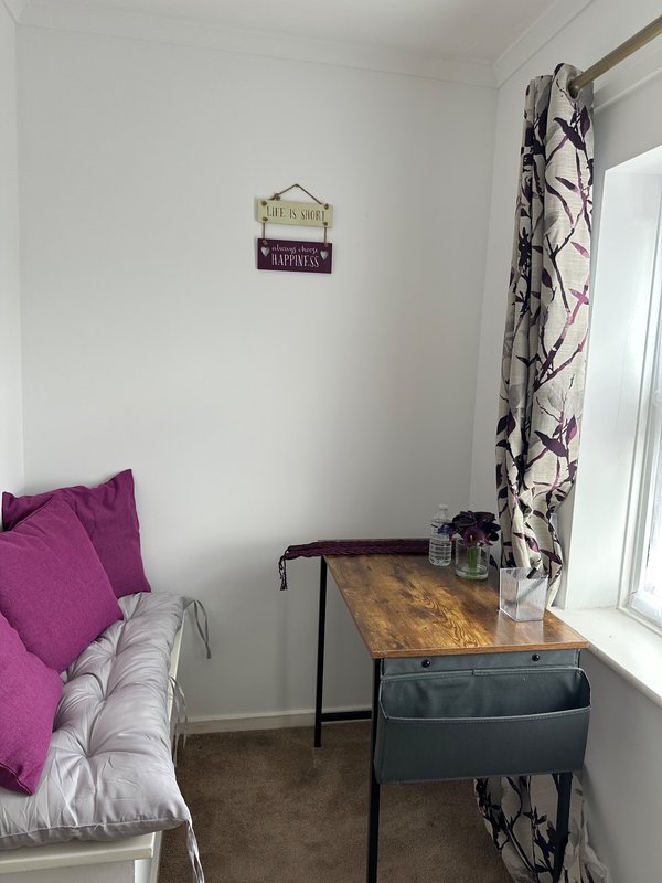 Bedroom with white walls, purple cushions on bed, wooden desk, and patterned curtains by window