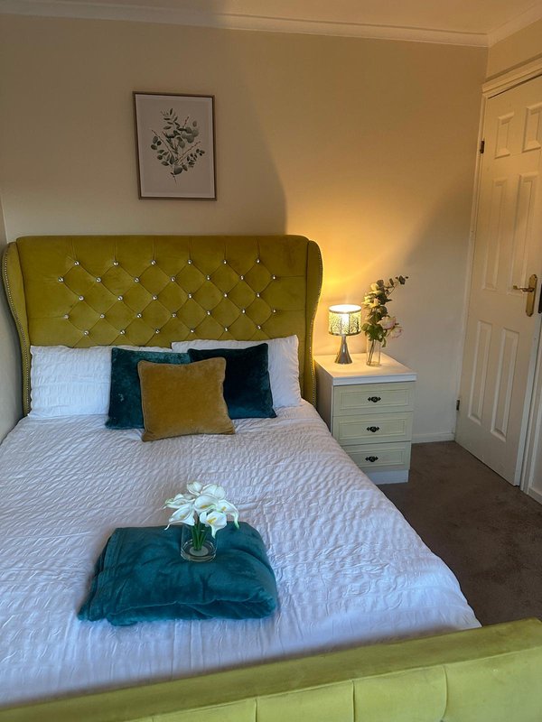 Bedroom with yellow upholstered headboard, white bedding, cushions, and bedside table with lamp