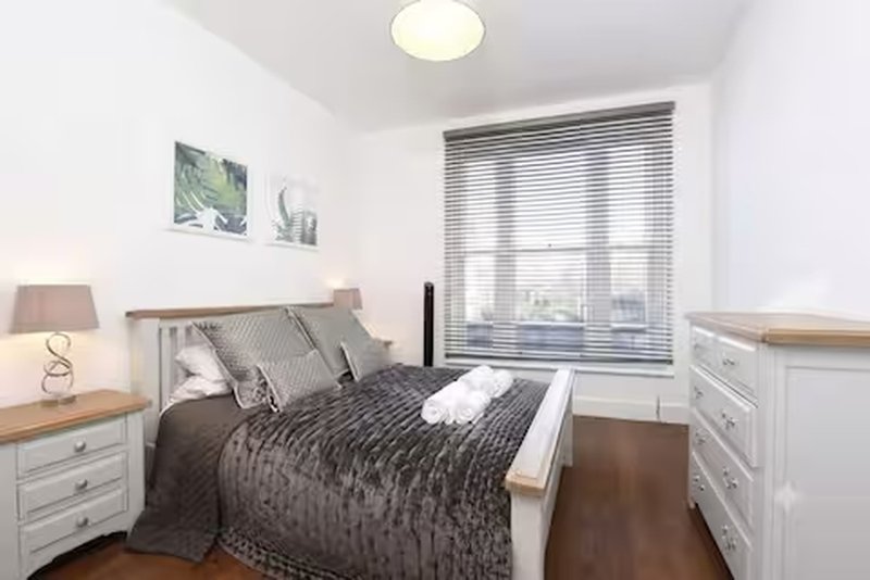 Modern bedroom with double bed, dark bedding, white furniture and large window with blinds