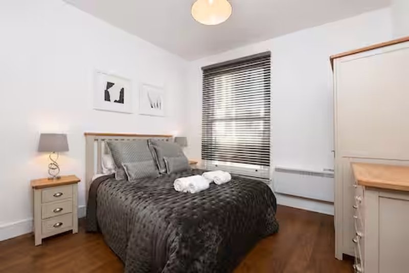 Modern bedroom with double bed, grey bedding, wooden nightstand, lamp and window blinds