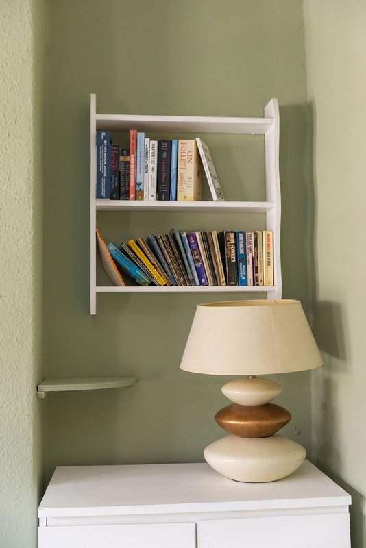 White wall-mounted bookshelf with books and table lamp with cream shade on white cabinet