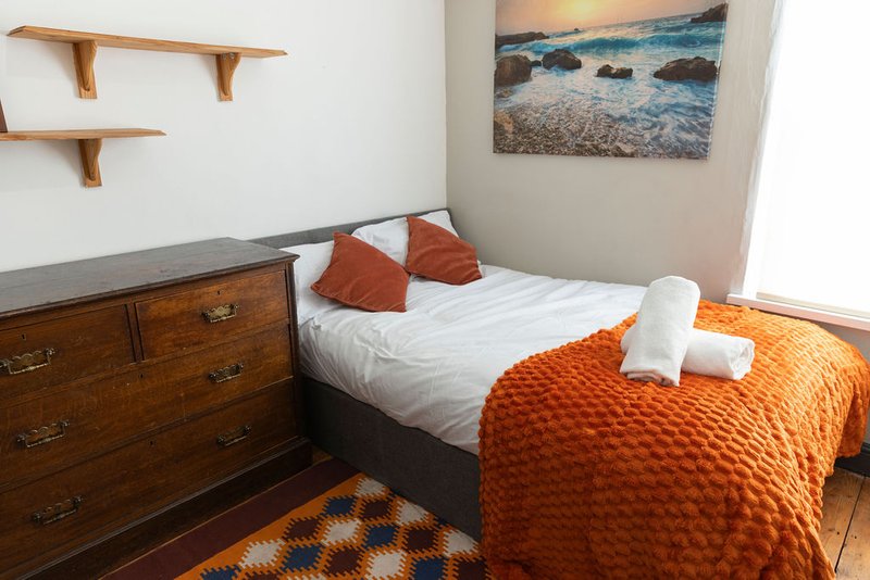 Bedroom with double bed, wooden dresser, orange bedding, wall shelves and beach artwork