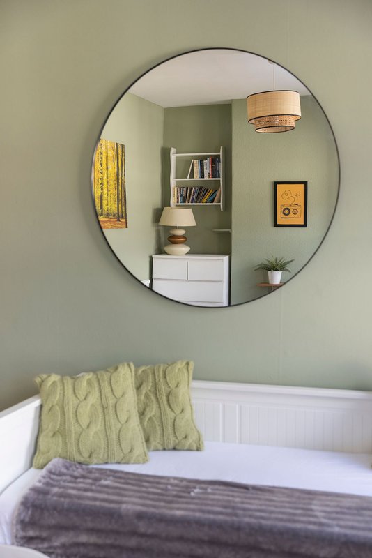 Bedroom with white panelled wall, round mirror, bed with green pillows and gray throw blanket