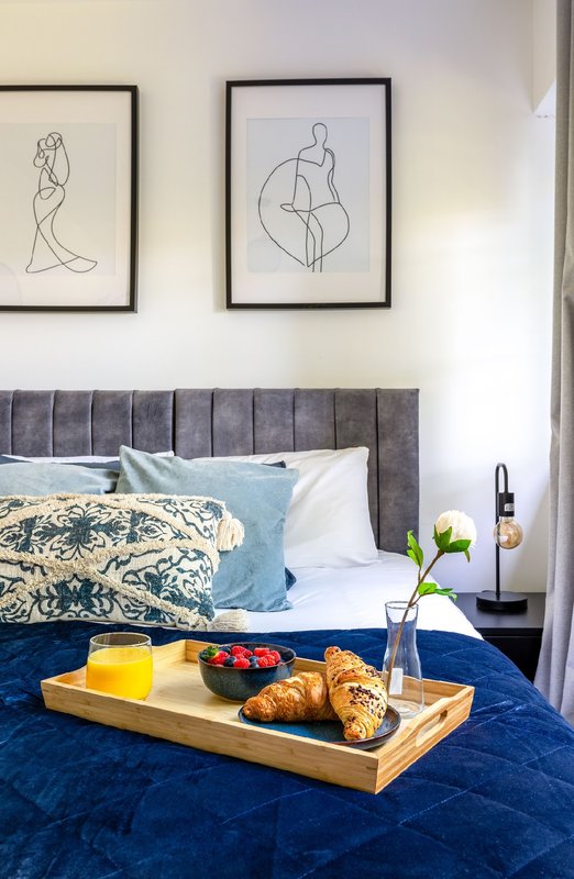 Bedroom with gray upholstered headboard, blue bedding, framed line art, and breakfast tray with pastries and flowers