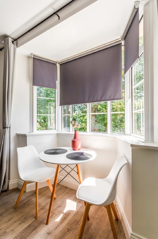 Bright dining nook with round white table, two white chairs, purple roller blinds and windows.