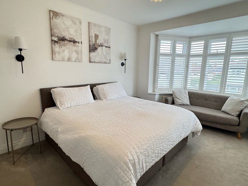 Modern bedroom with white bedding, grey headboard, wooden side table, wall art and large shuttered windows