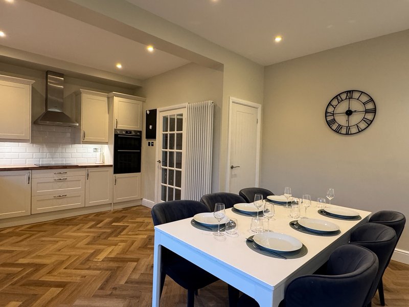 Open-plan kitchen and dining area with cream cabinetry, dining table set for six, and wall clock