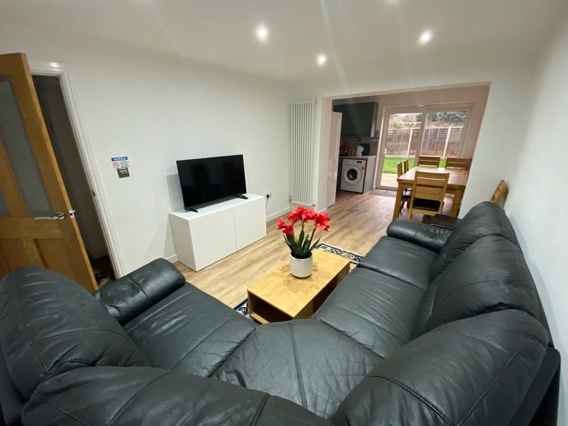 Living room with black leather sofa, TV, wooden table, flowers and kitchen area visible