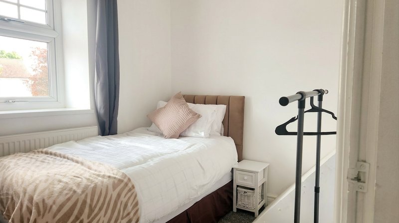 Bedroom with single bed, beige bedding, pink cushion, wooden headboard, and window with curtains