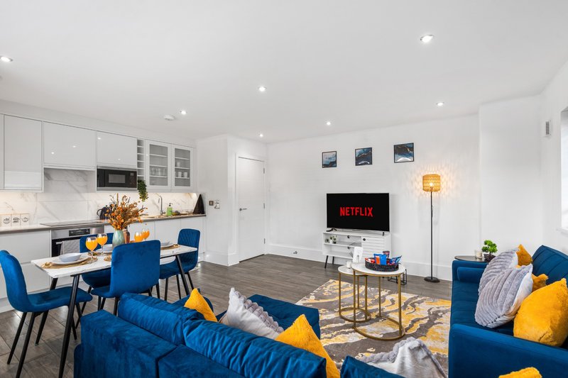 Open-plan living area with blue sofa, dining table, kitchen, and TV mounted on wall