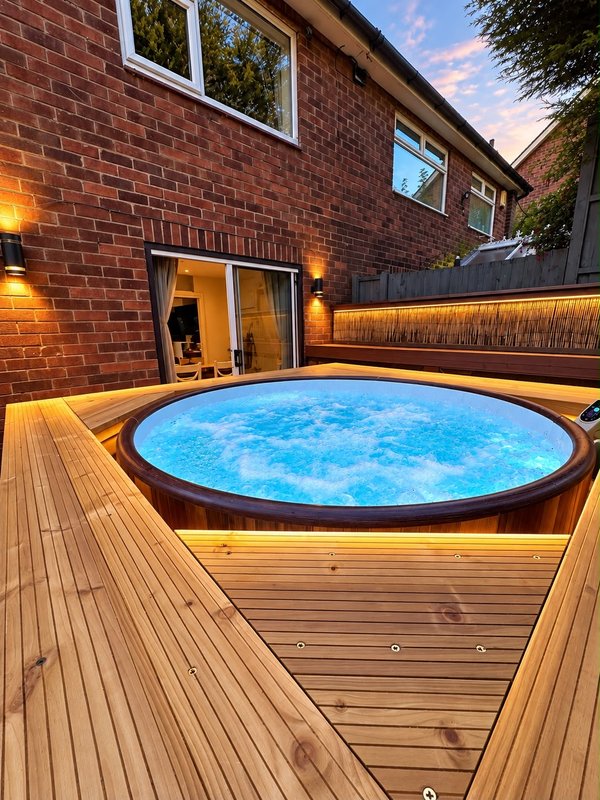 Modern backyard with illuminated hot tub, wooden decking, brick house and patio lighting