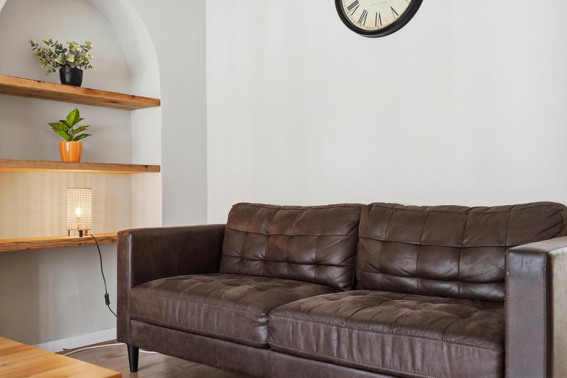 Living room with brown leather sofa, wooden shelves with plants, and wall clock