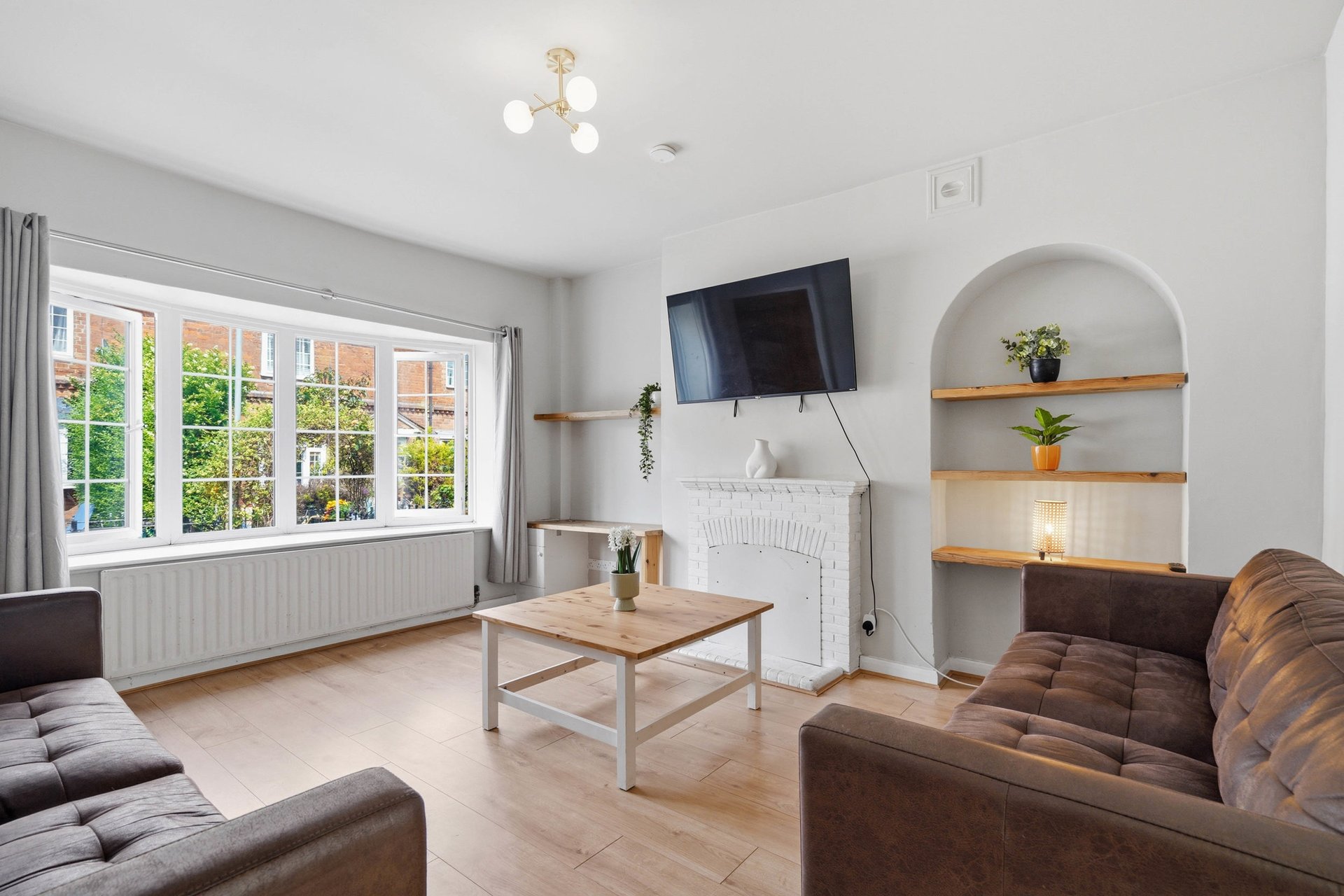 Bright living room with brown leather sofas, wooden coffee table, fireplace, shelving, and garden views