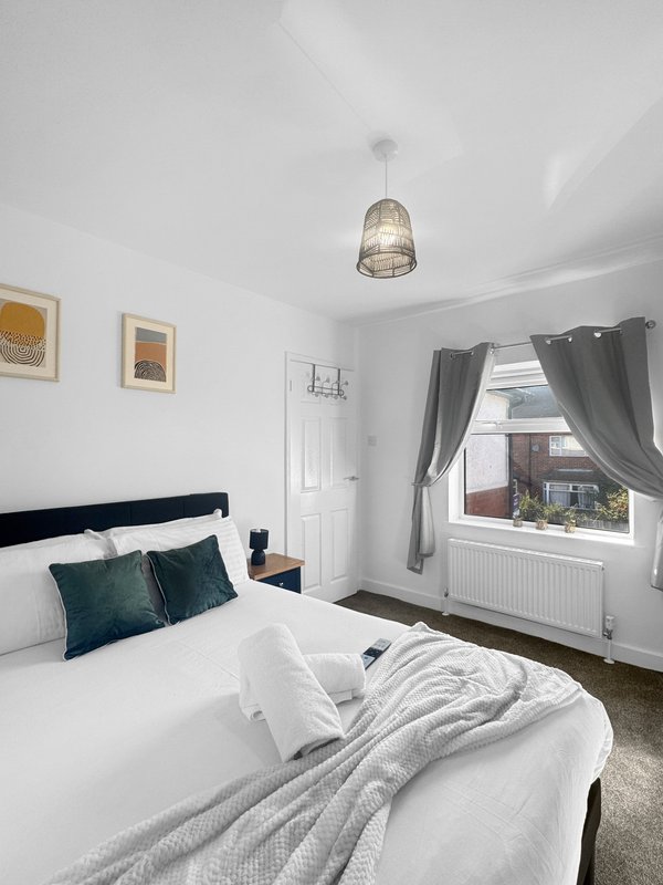 Bright bedroom with double bed, dark headboard, teal pillows, pendant light and window with curtains