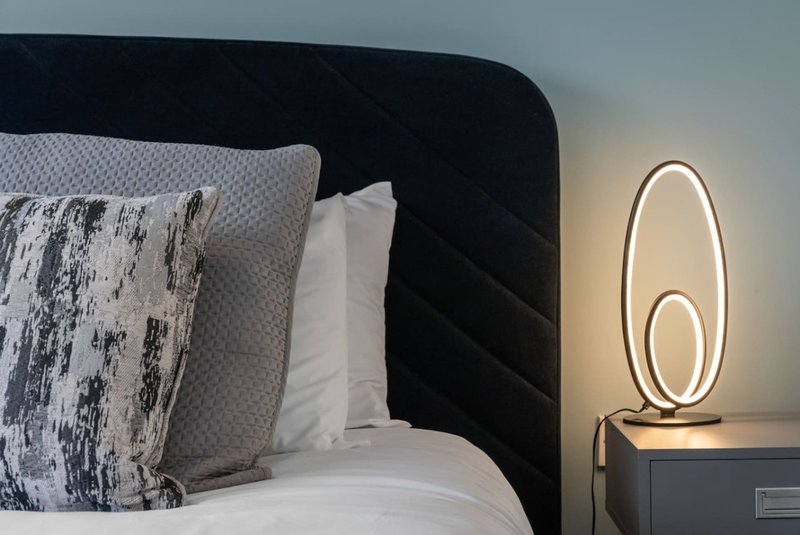 Modern bedroom with black upholstered headboard, patterned cushions, white bedding, and oval neon bedside lamp
