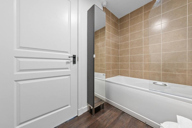 Modern bathroom with white door, beige tile walls, white bathtub and dark wood flooring