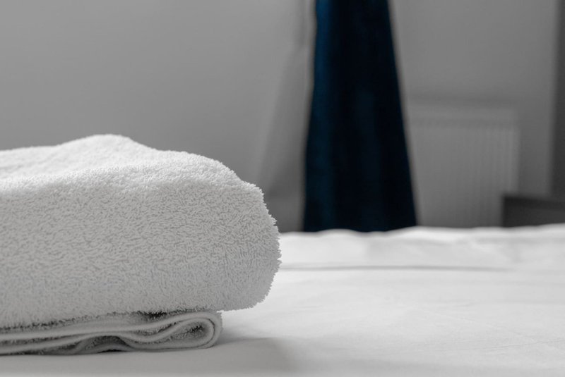Rolled white towel on bed with dark clothing hanging in background