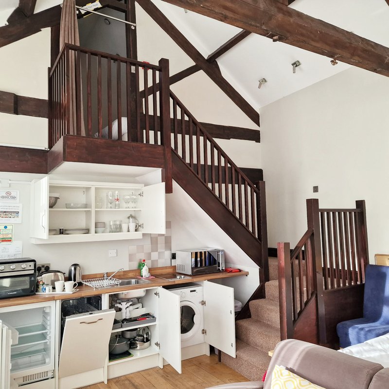 Open-plan studio with kitchenette, wooden beamed ceiling, and wooden staircase to mezzanine
