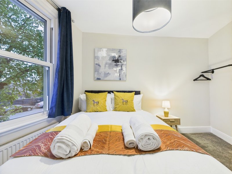 Bright bedroom with double bed, yellow pillows, rolled towels, and large window with blue curtains