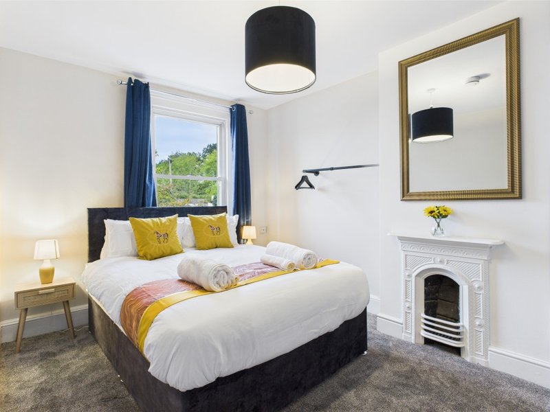 Bright bedroom with double bed, yellow cushions, fireplace, mirror and window with blue curtains