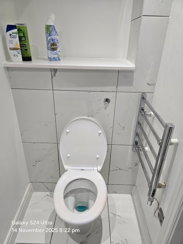 Bathroom toilet with white ceramic tiles, shelf with toiletries, and heated towel rack