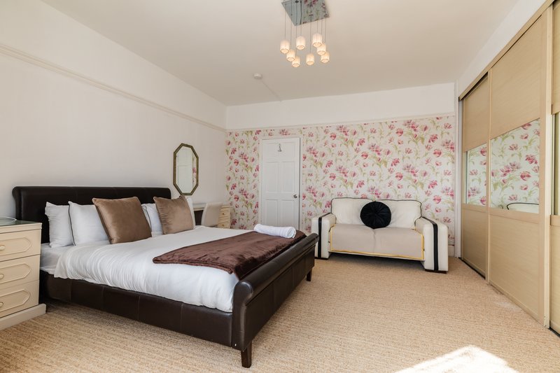 Spacious bedroom with dark leather bed, white bedding, floral wallpaper, cream carpet and pendant lighting