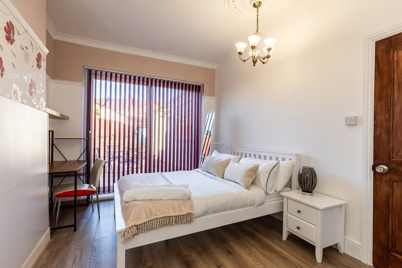 Bright bedroom with double bed, white nightstand, desk, and vertical blinds by patio door