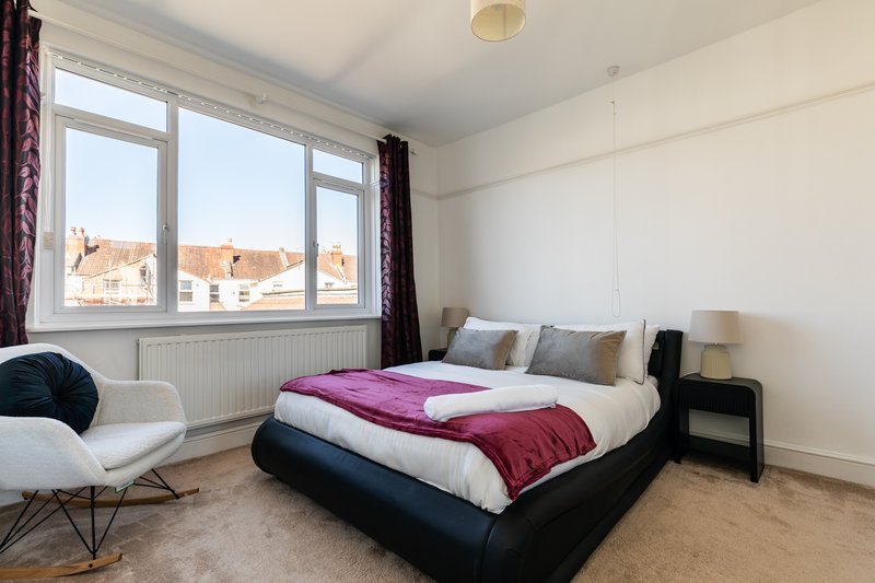 Bright bedroom with double bed, burgundy throw, white chair, and large windows overlooking street