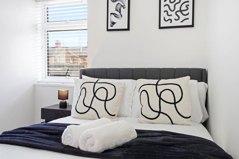 Modern bedroom with dark upholstered headboard, patterned cushions, rolled towels and city view windows