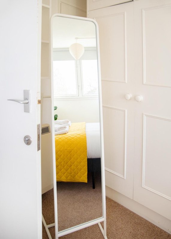 Bedroom viewed through open door, showing full-length mirror, yellow bedding, and cream-colored walls