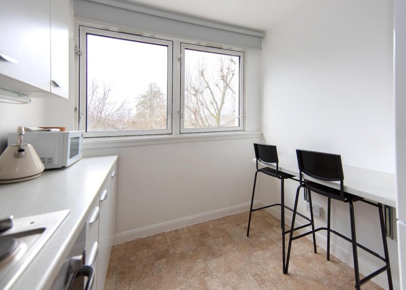 Modern kitchen with white counters, stainless steel appliances, two bar stools, and window overlooking bare trees