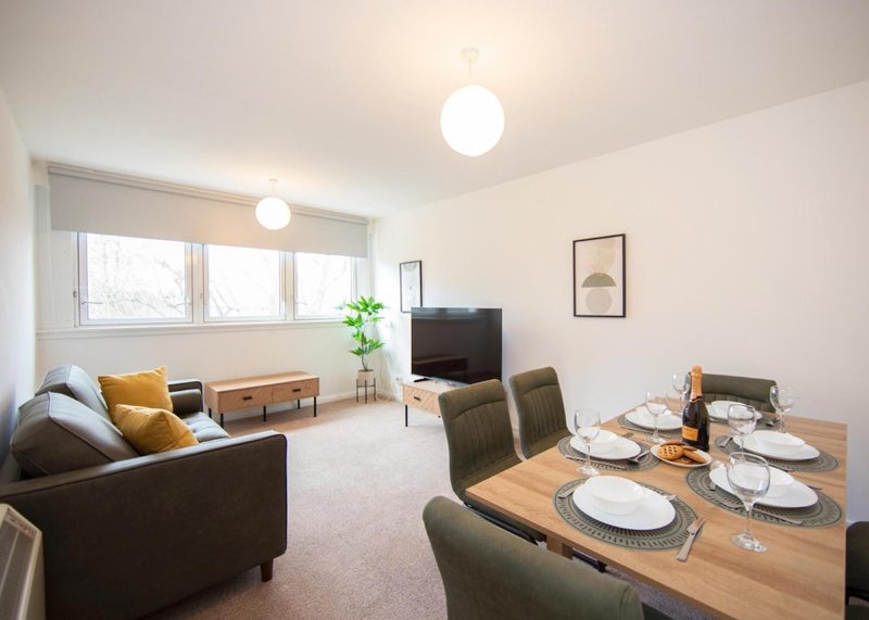Open-plan living and dining area with sofa, dining table set for four, TV, and large windows